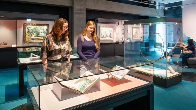 Two female students look at two books in a glass case. A large glass case with a brass horn is displayed to their right and paintings are shown in the glass cases behind them. 