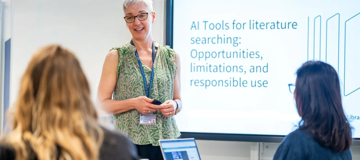 Presenter leading an in-person academic workshop at a university library. Two attendees are seated in the foreground with a laptop on the table, listening to the talk in a  training room setting.