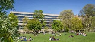 People relaxing on a sunny day on the Meadows, with the Main Library in the background.