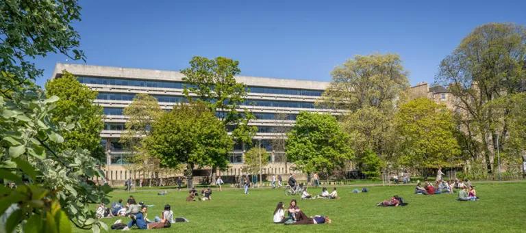 People relaxing on a sunny day on the Meadows, with the Main Library in the background.