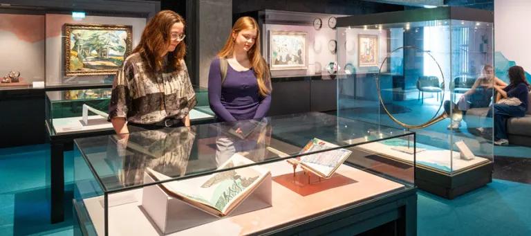 Two people observe glass display cases containing books and artifacts in The Beathing Room, Main Library with framed artwork on dark walls.