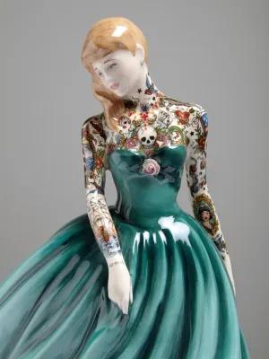 A porcelain figurine with a teal dress and colourful tattoos along the arms, neck and chest.